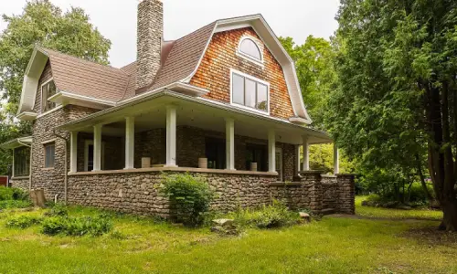 Stone house with wraparound porch and greenery.